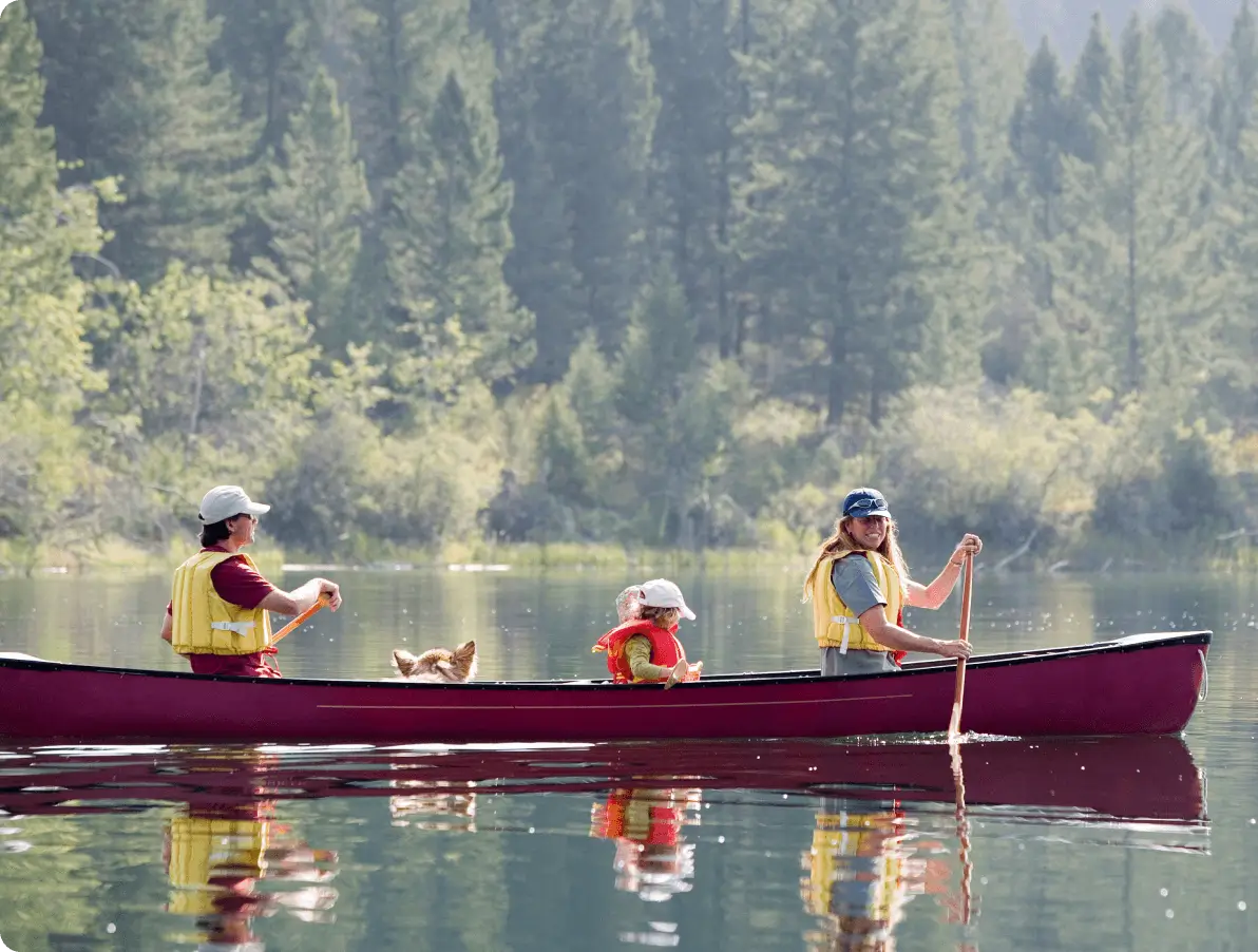 A family with a dog paddles a red canoe on a calm lake surrounded by a forested landscape.