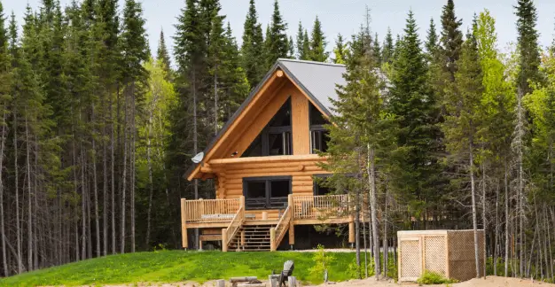 A wooden A-frame cabin stands near a tranquil lake, surrounded by pine trees, with its reflection visible in the water.