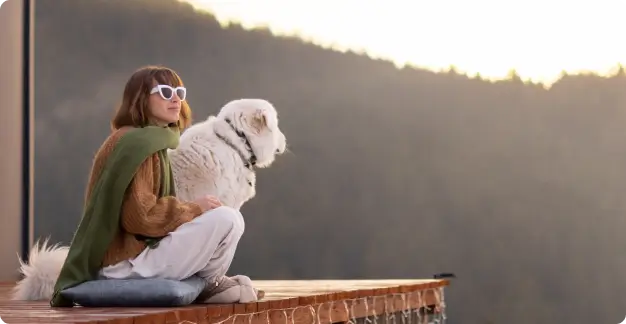 A person wearing sunglasses and holding a white dog sits on a platform, overlooking a mountainous landscape at sunrise.